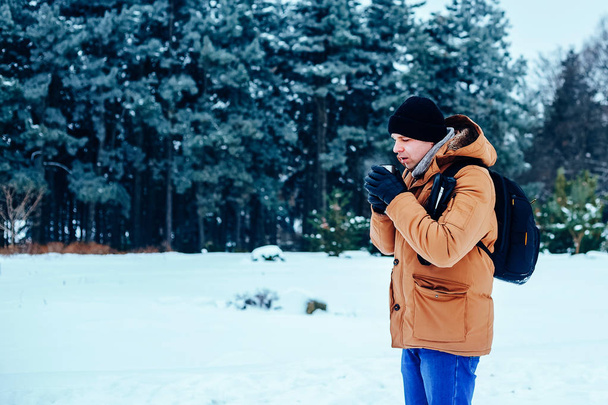 un giovanotto in giacca rossa con uno zaino in piedi sulla neve e con in mano un thermos e una tazza di tè. Foresta invernale
. - Foto, immagini
