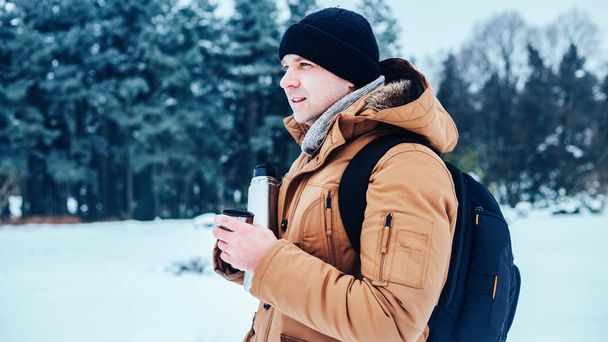 雪の上に立っていると、ポットと紅茶のマグカップを保持しているバックパックと赤いジャケットの若い男。冬の森. - 写真・画像
