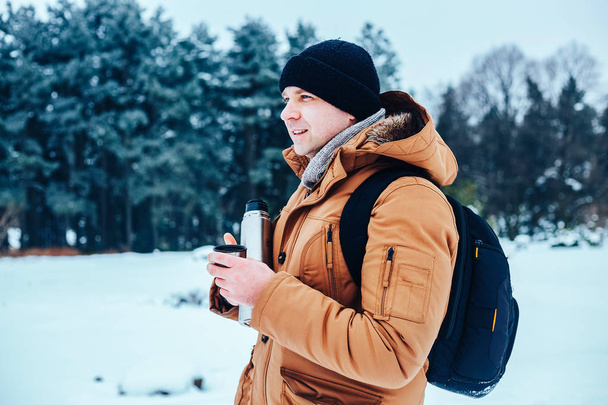 молодий чоловік в червоній куртці з рюкзаком, що стоїть на снігу і тримає термос і чашку чаю. Зимовий ліс
. - Фото, зображення