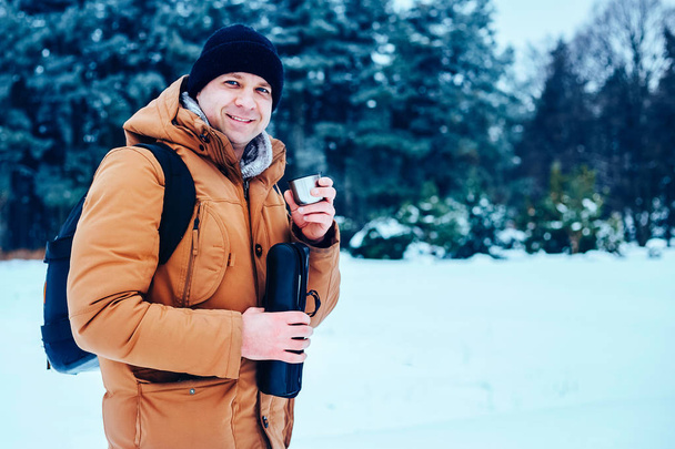 a young man in a red jacket with a backpack standing on the snow and holding a thermos and a mug of tea. Winter forest. - Foto, immagini