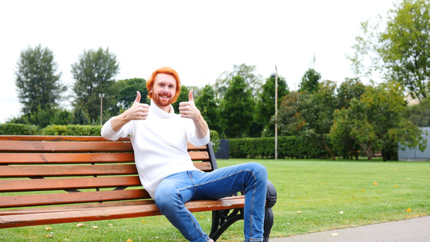 Daumen hoch von Mann, der auf Bank im Park sitzt, rote Haare und Bart - Foto, Bild