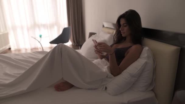 Girl lying in bed in the morning. just woke up and looks into the phone. White apartment, brunette - Video, Çekim