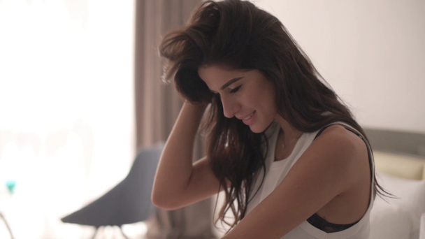 girl with dark hair sitting on the bed in the morning, cute smiles into the camera. White bedroom, Sun behind the window - Footage, Video