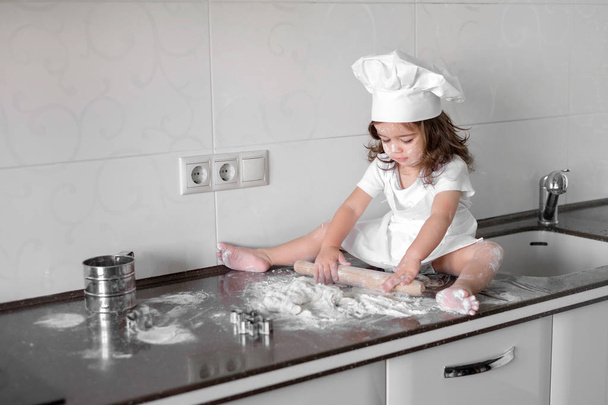 Little smiling baby girl baker in white cook hat and apron kneads a dough on tle kitchen - Fotografie, Obrázek