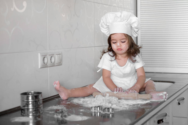 Little smiling baby girl baker in white cook hat and apron kneads a dough on tle kitchen - Фото, зображення