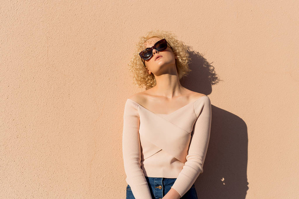 beautiful young girl with curls in sunglasses stands near the wall in sunlight at sunset - Foto, afbeelding