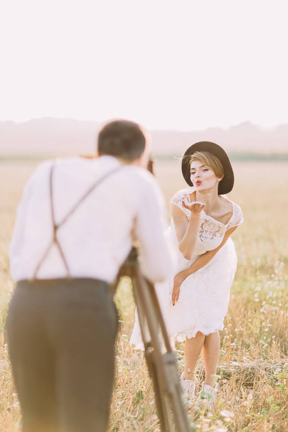 The blurred back view of the groom taking photos of the bride sending kisses in the sunny field. - Φωτογραφία, εικόνα