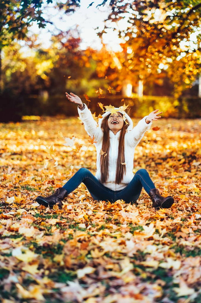 Gelukkig jonge vrouw in park op zonnige herfstdag, lachen, spelen bladeren. Vrolijke mooi meisje in witte trui tijdens herfst seizoen in het park - Foto, afbeelding