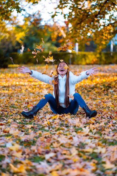Happy young woman in park on sunny autumn day, laughing, playing leaves. Cheerful beautiful girl in white sweater during autumn season in the park - Photo, Image