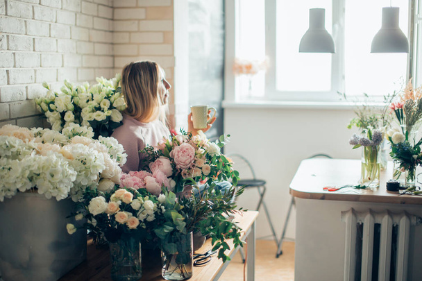 Florist at work. woman in process of making bouquet, holds hand cup of coffee - Foto, afbeelding
