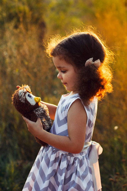 Little cute curly girl hugd a fluffy toy owl. Toddler girl play with sweet doll. A beautiful sunlight, warm colours. Autumn time - Photo, Image