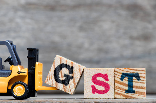 Жовтий пластиковий навантажувач тримає літеру G для повного слова GST (погашення податку на товари та послуги) на фоні деревини
 - Фото, зображення