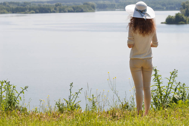 Een roodharige jonge vrouw in een hoed staat op een hoge oever van het meer en in de verte kijkt. - Foto, afbeelding