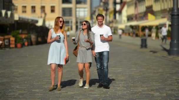 Tres amigos felices tomando café mientras caminan por la ciudad. Retrato en altura de dos niñas y un niño con bebidas en las manos
. - Metraje, vídeo