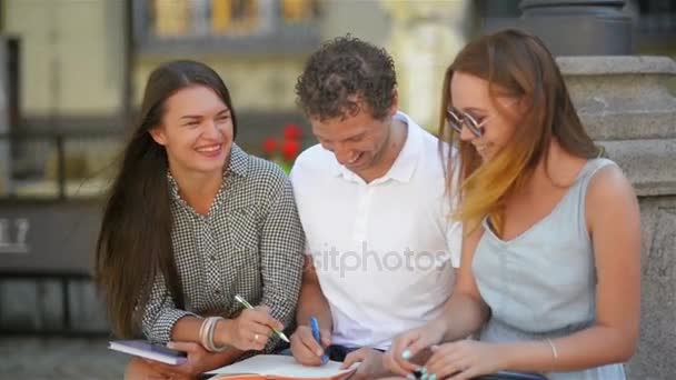 Drei Studenten lernen draußen und lachen gemeinsam an einem warmen, sonnigen Tag in der Stadt. zwei Mädchen und ein Junge mit Notizen, Kugelschreibern und Tabletten für die Prüfung. - Filmmaterial, Video