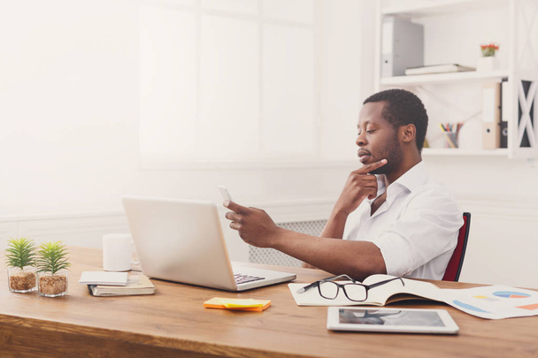Задумчивый афро-американский бизнесмен пишет смс по телефону во время работы над ноутбуком
 - Фото, изображение
