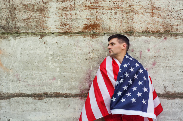jonge kerel verpakt in Amerikaanse vlag van de Verenigde Staten ten opzichte van de betonnen muur. expressieve emoties kijken - Foto, afbeelding