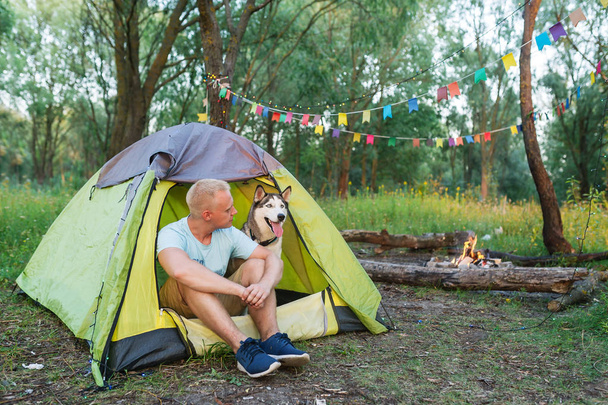 Cara com um cão husky em férias sentado na natureza ao lado da tenda à noite ao pôr do sol
 - Foto, Imagem