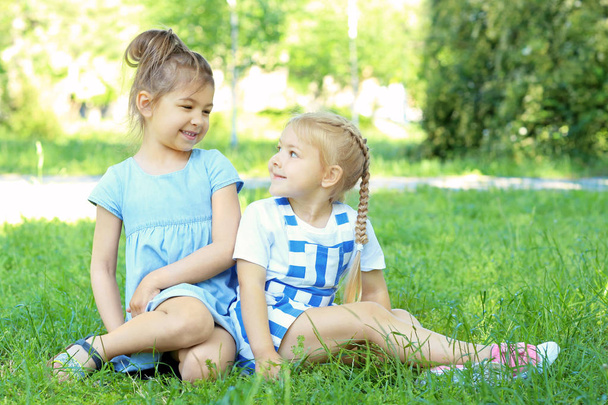 Schattige kleine meisjes zitten op groen gras in park - Foto, afbeelding