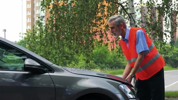 Starší muž nosí výstražná vesta, otevře kapotu a kontroly motoru automobilu - budovy a strom v pozadí - Záběry, video