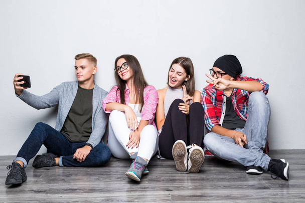 Grupo de estudantes bonitos em roupas casuais fazendo selfie usando um smartphone e sorrindo enquanto sentados juntos no chão
 - Foto, Imagem