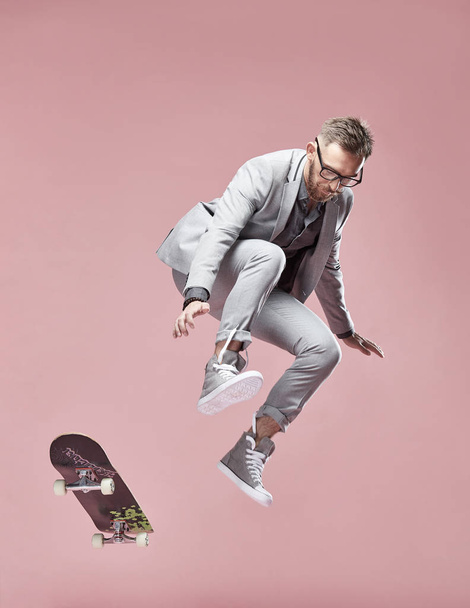 Young handsome serious man with glasses, brown hair and beard, wearing light grey suit and sneakers, jumping with the skateboard on light pink background  - Fotó, kép