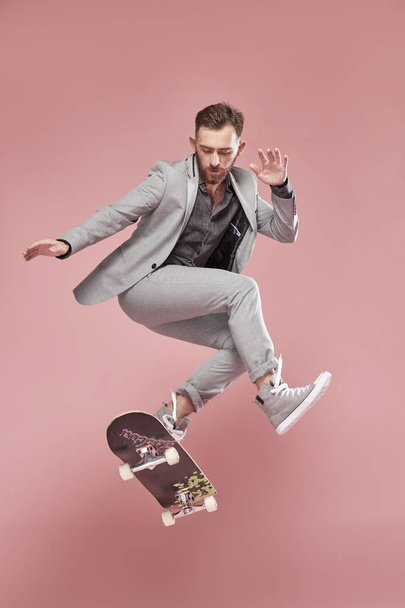 Young handsome man with brown hair and beard, wearing light grey suit and sneakers, jumping with the skateboard on light pink background  - Foto, Imagem