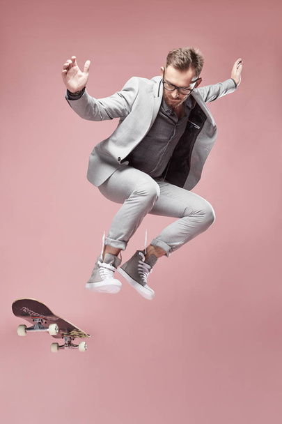 Young handsome serious man with glasses, brown hair and beard, wearing light grey suit and sneakers, jumping with the skateboard and flying on light pink background  - Фото, зображення