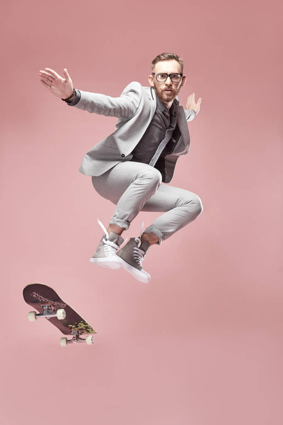 Young handsome serious man with glasses, brown hair and beard, wearing light grey suit and sneakers, jumping with the skateboard and flying in the air on light pink background  - Fotografie, Obrázek