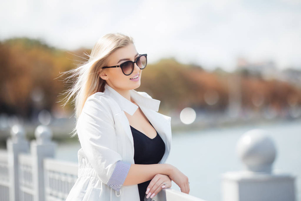Όμορφη νεαρή γυναίκα που θέτουν ως μοντέλο στέκεται στο ανάχωμα κοντά στη θάλασσα ή τον ωκεανό. Κυρία φορούν casual και γυαλιά ηλίου. - Φωτογραφία, εικόνα