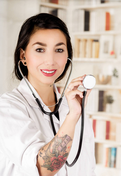 Beautiful smiling tattooed young doctor holding the chest part of the stethoscope in her hand, in office background - Fotoğraf, Görsel