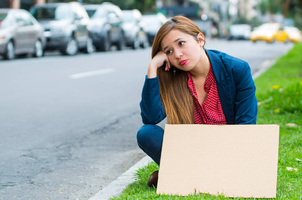 Young woman wearing casual clothes sitting outdoors holding cardboard poster, protesting concept - Photo, Image
