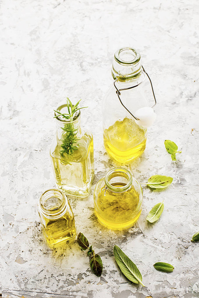 Four bottles of different sizes with vegetable oil, sunflower and olive on a light marble background with the addition of pepper, sage leaves and basil for kitchen and cosmetic use. Selective focus. - Φωτογραφία, εικόνα