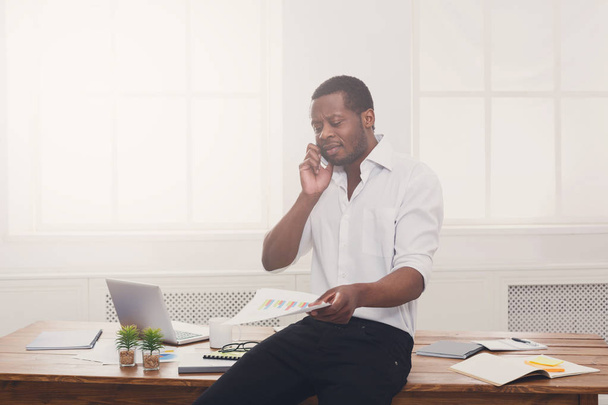 junger schwarzer Geschäftsmann liest Dokumente und spricht auf seinem Handy in einem modernen weißen Büro - Foto, Bild