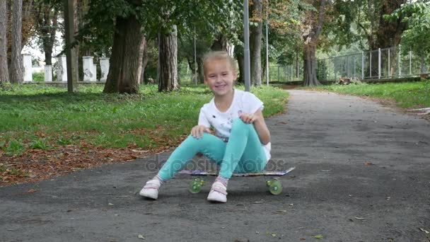 Een meisje zit op het skateboard en van links naar rechts rijden. Kind op zoek naar de camera terwijl zittend op het skateboard.  - Video