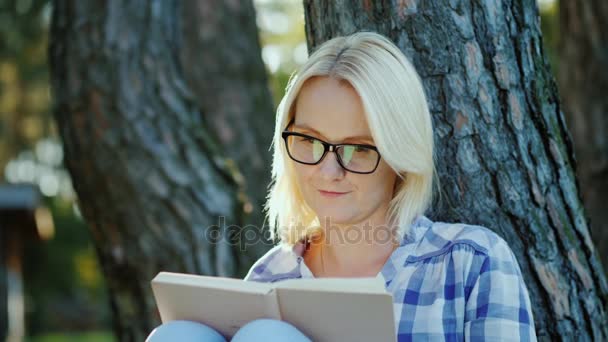 A blonde young woman in glasses reads a book in the park. Sits near a tree, beautiful light before sunset - Video