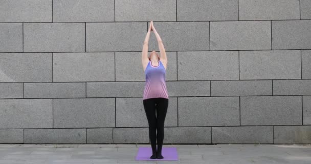 Woman practices yoga fitness exercise on pink mat in the city on grey urban background - Footage, Video