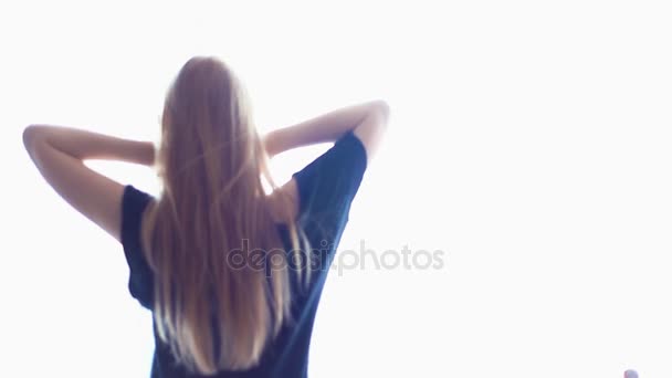 Joven chica rubia acaricia el cabello y se estira delante de la ventana brillante
 - Metraje, vídeo