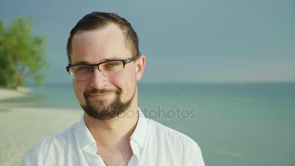 Portrét mladého muže na tropické pláži. V jednoduché tričko usměje se a dívá do kamery. Nosí brýle a vousy - Záběry, video