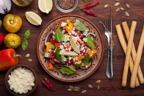 Salad dish made of tomatoes, peppers, cucumbers, purple onions, basil leaves, cottage cheese, olive oil and lime juice on a dark wooden background. A useful dish. Healthy eating, balanced nutrition - Photo, Image