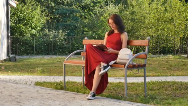 Zeki kız öğrenci parkta bankta oturmuş ve bir şey bir defterde yazılı Eyesglasses ve kırmızı uzun elbise. - Video, Çekim