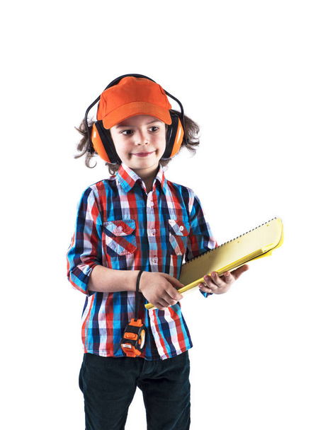 Little curly boy builder in a orange cap holds a workbook and roulette . White background - Zdjęcie, obraz