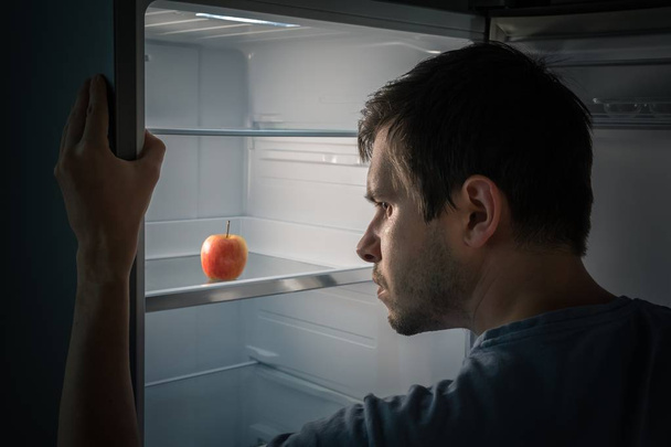 Hongerige man is op zoek naar voedsel in de koelkast bij nacht. Alleen apple is - Foto, afbeelding