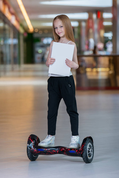 Happy and smiling girl rides on mini segway at trading mall. Teenager riding on hover board or gyroscooter and holding blank sheet. - Фото, изображение