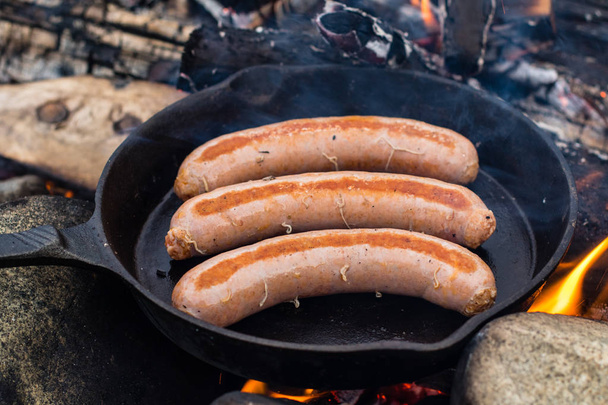 Cooking sausages in cast iron skillet on campfire while camping. Good and positive campfire food. - Photo, Image