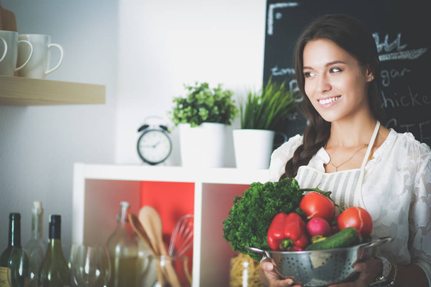 Smiling young woman holding vegetables standing in kitchen - Foto, Bild