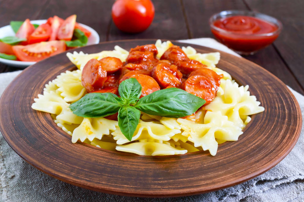 Farfalle pasta in a ceramic bowl, serve with ketchup, fresh tomatoes, sausage in tomato sauce on a dark wooden background. - Photo, Image