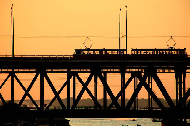 Silhouetten einer zweistufigen Brücke und 2 Straßenbahnen vor dem Hintergrund des Sonnenuntergangs - Foto, Bild