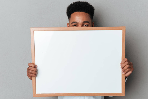 Young afro american man hiding behind blank board - 写真・画像