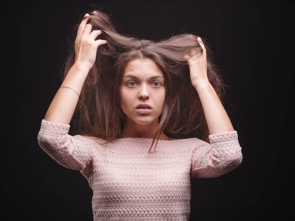 Shocked female pulling her sick hair on a black background. Gorgeous woman unhappy with her hair. Hair routine concept. - Фото, изображение